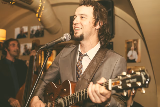 Gitarist pjeva uz mikrofon i svira gitaru u svođenoj birtiji